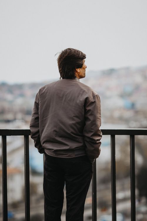 Free Senior man in jacket stands by railing, contemplating city skyline, captured in a vertical shot. Stock Photo