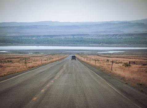 A solitary car travels a deserted highway lined by brown fields, approaching distant mountains.