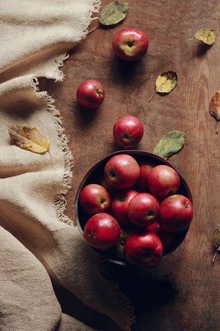 A top view of red apples in a bowl amidst autumn leaves on a rustic wooden table.