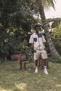 Young man in casual attire with vintage TV by tropical garden backdrop.