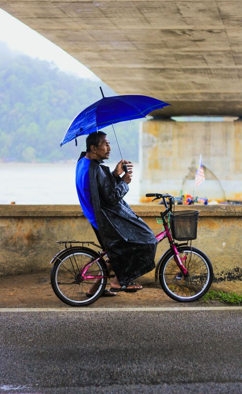 Free Elderly man rides bicycle under a bridge, holding blue umbrella in rainy weather. Stock Photo