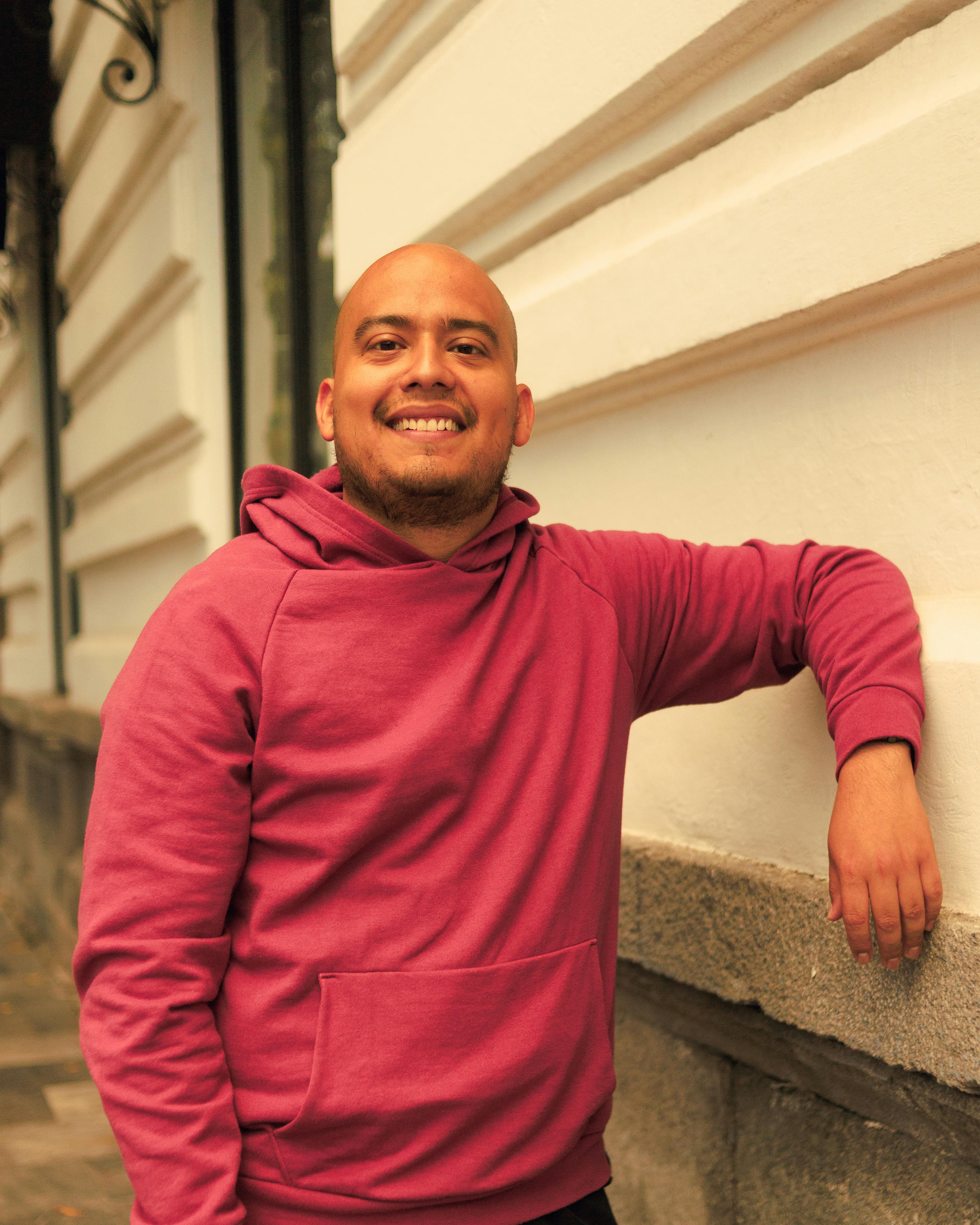 Bald man smiling confidently in a pink hoodie against urban architecture.