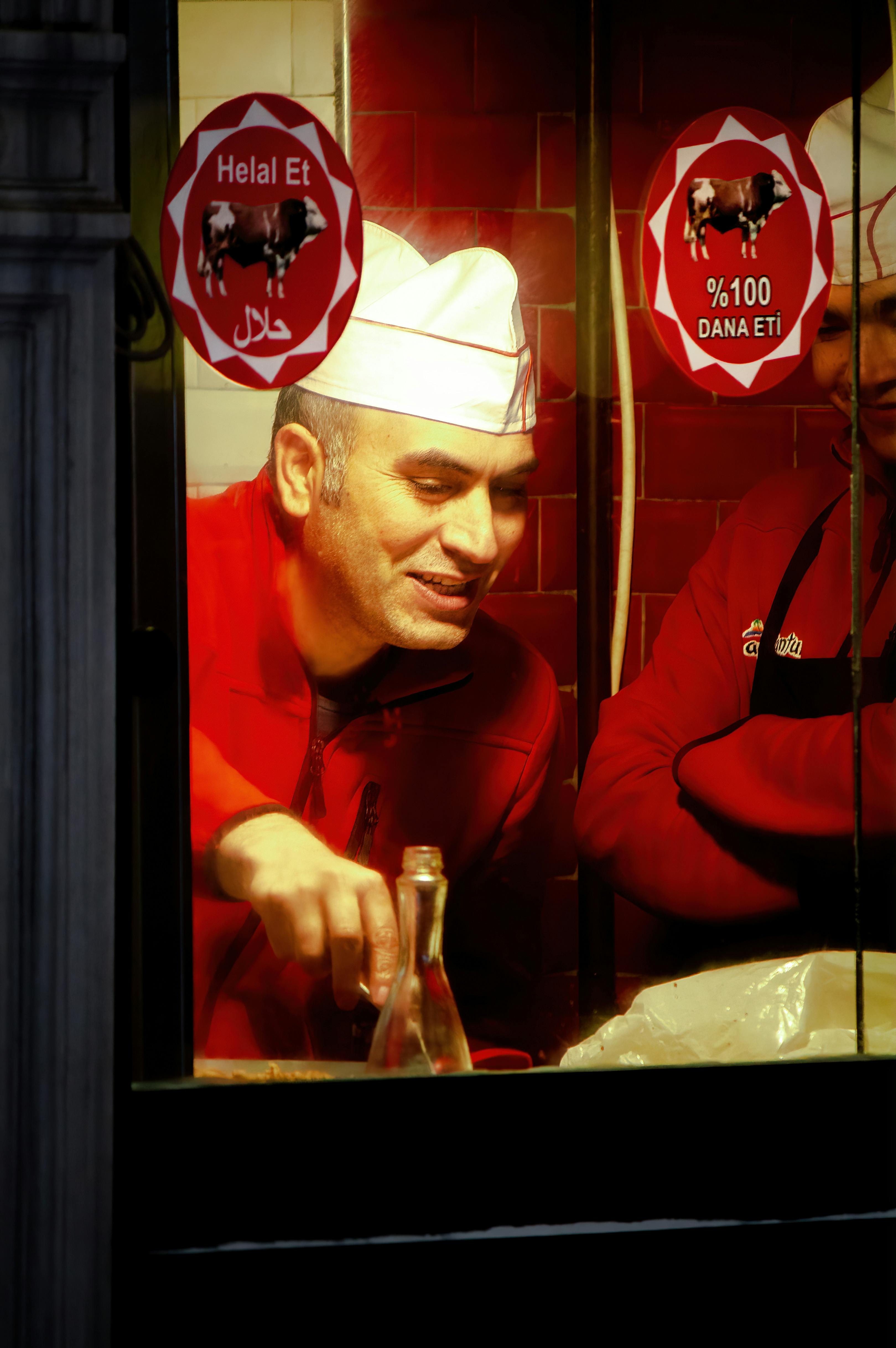 Two male chefs in red uniforms preparing meat in a kitchen viewed through a window