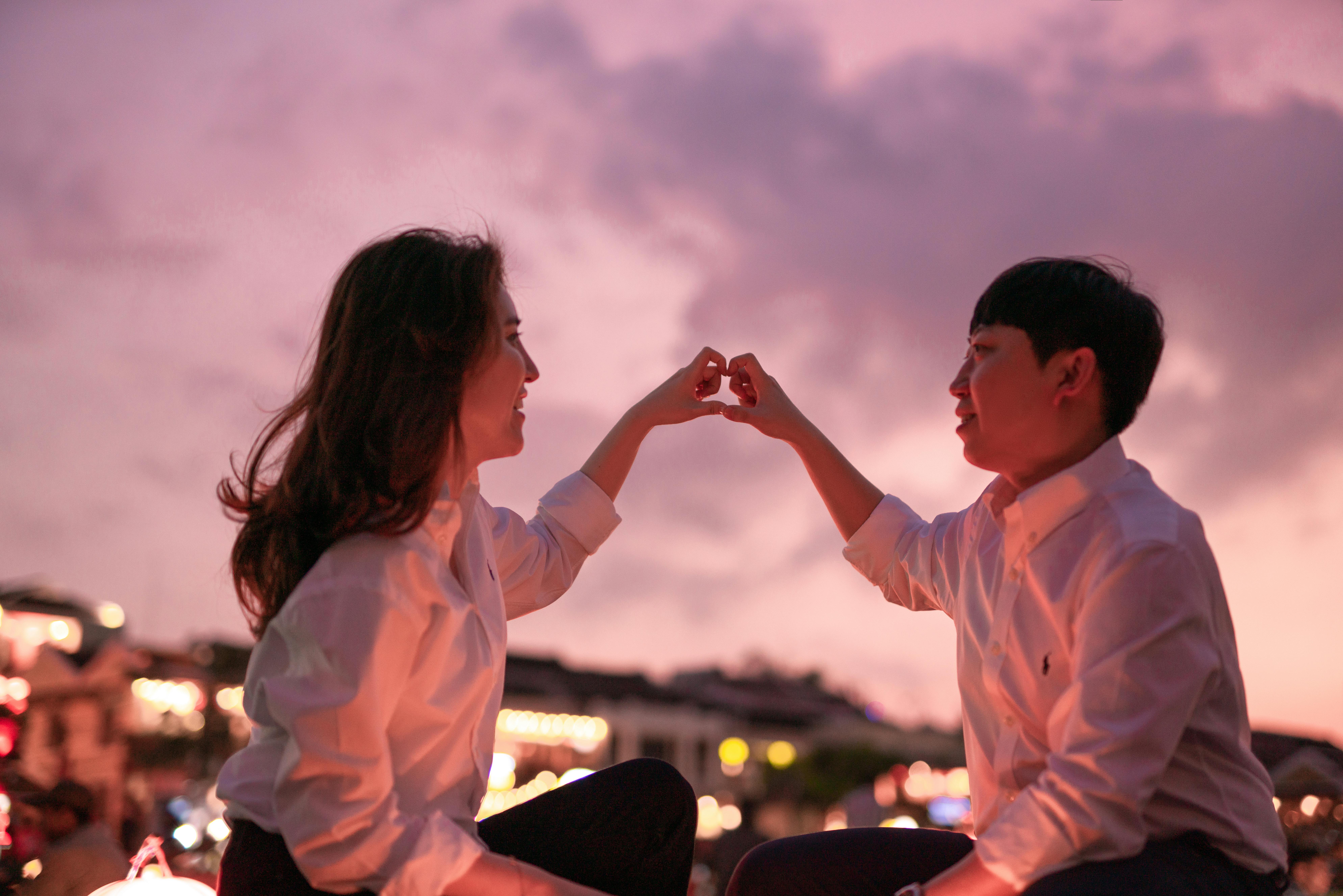 A couple in white shirts forms a heart shape with their hands against a vibrant sunset sky.