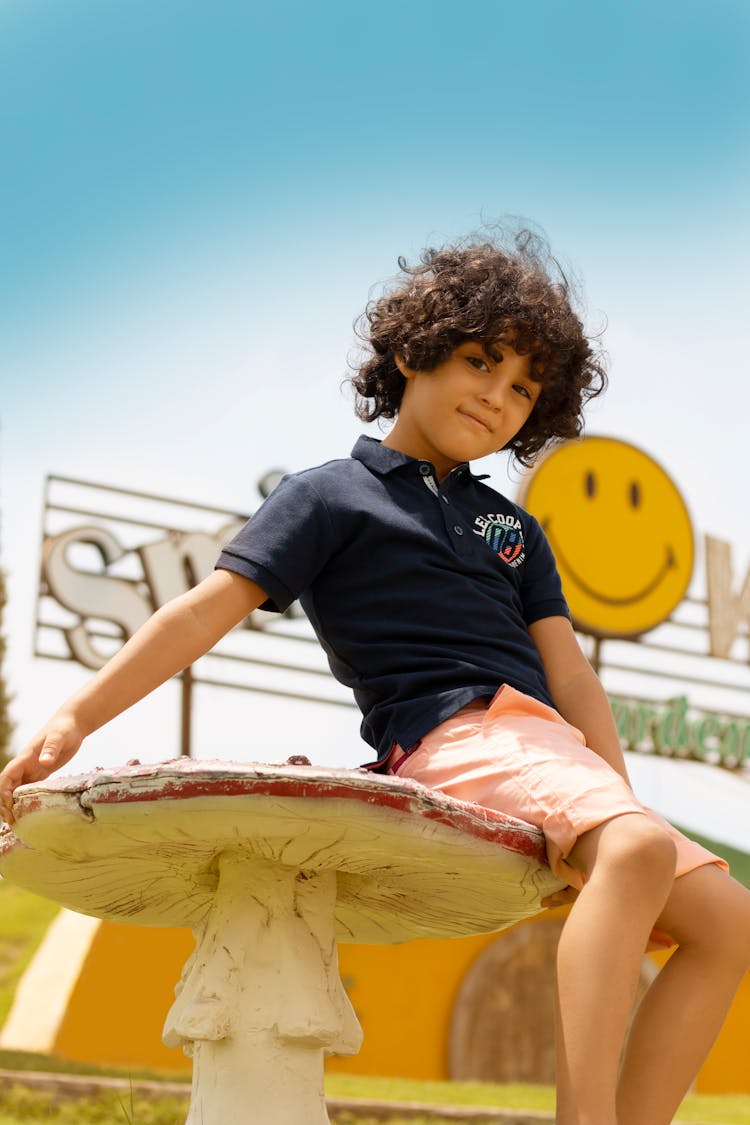 Cute Little Boy In Blue Shirt Sitting On A Mushroom Statue