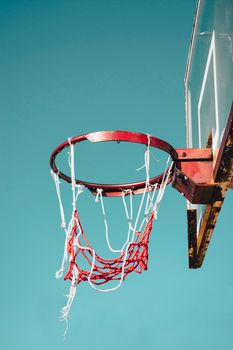 A rusty outdoor basketball hoop with a weathered, broken net against a clear blue sky.