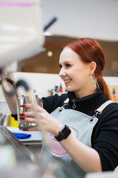 A cheerful barista with red hair froths milk at a cafe, showcasing professional coffee-making skills.