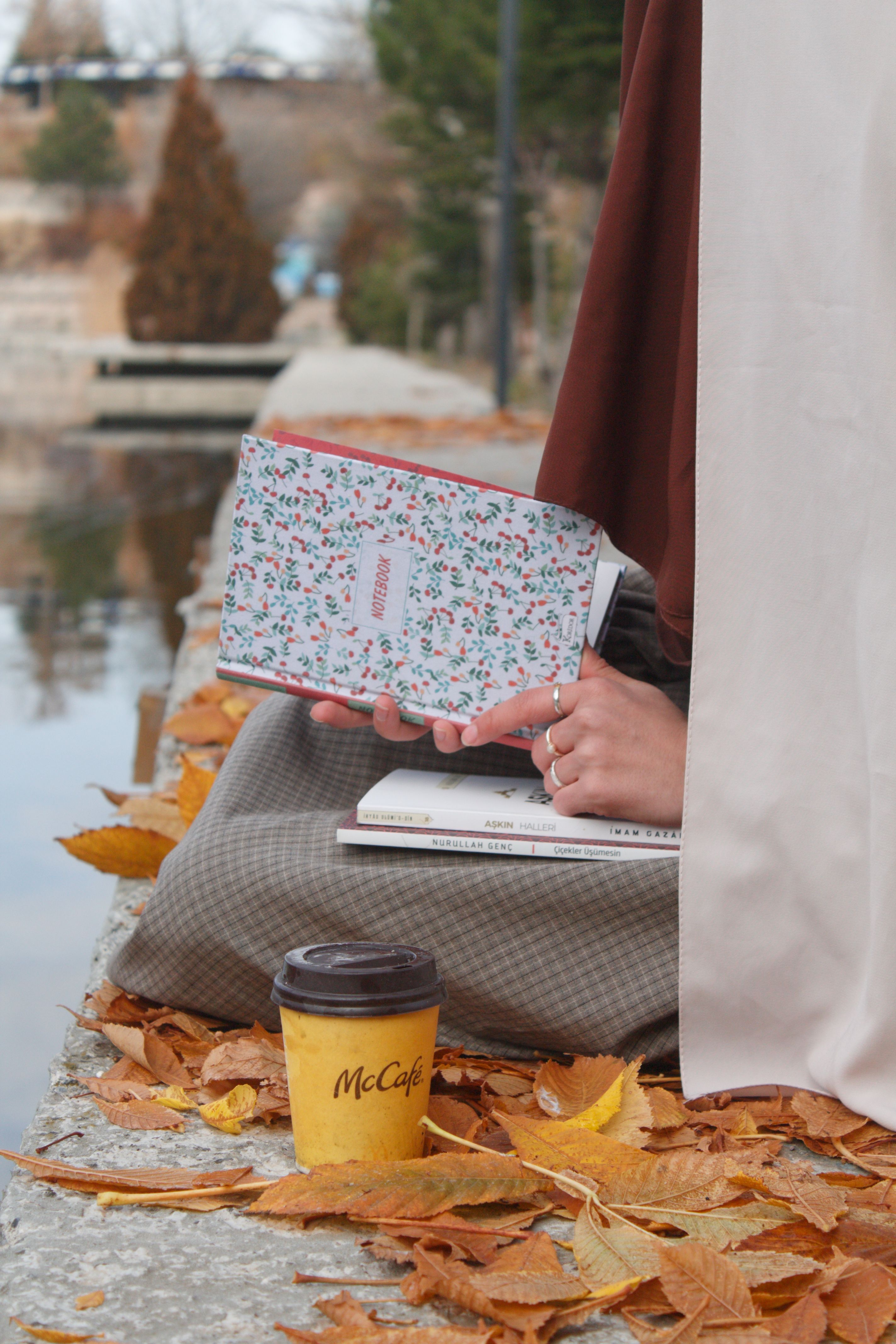 A person holding a floral book and coffee cup surrounded by fallen leaves by the lake.