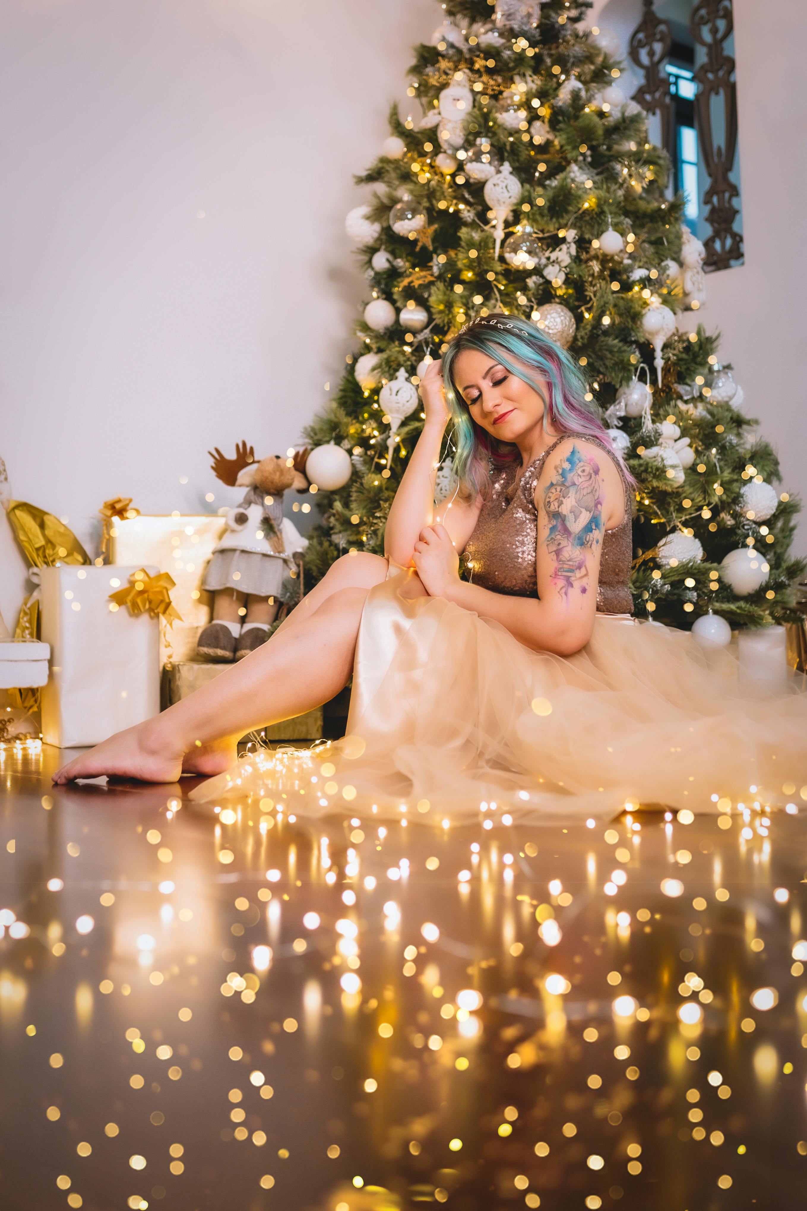 Woman with colored hair and tattoos sitting by a Christmas tree amidst twinkling lights.