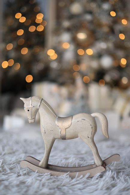 Wooden rocking horse in focus, with festive blurred Christmas tree and lights in background.