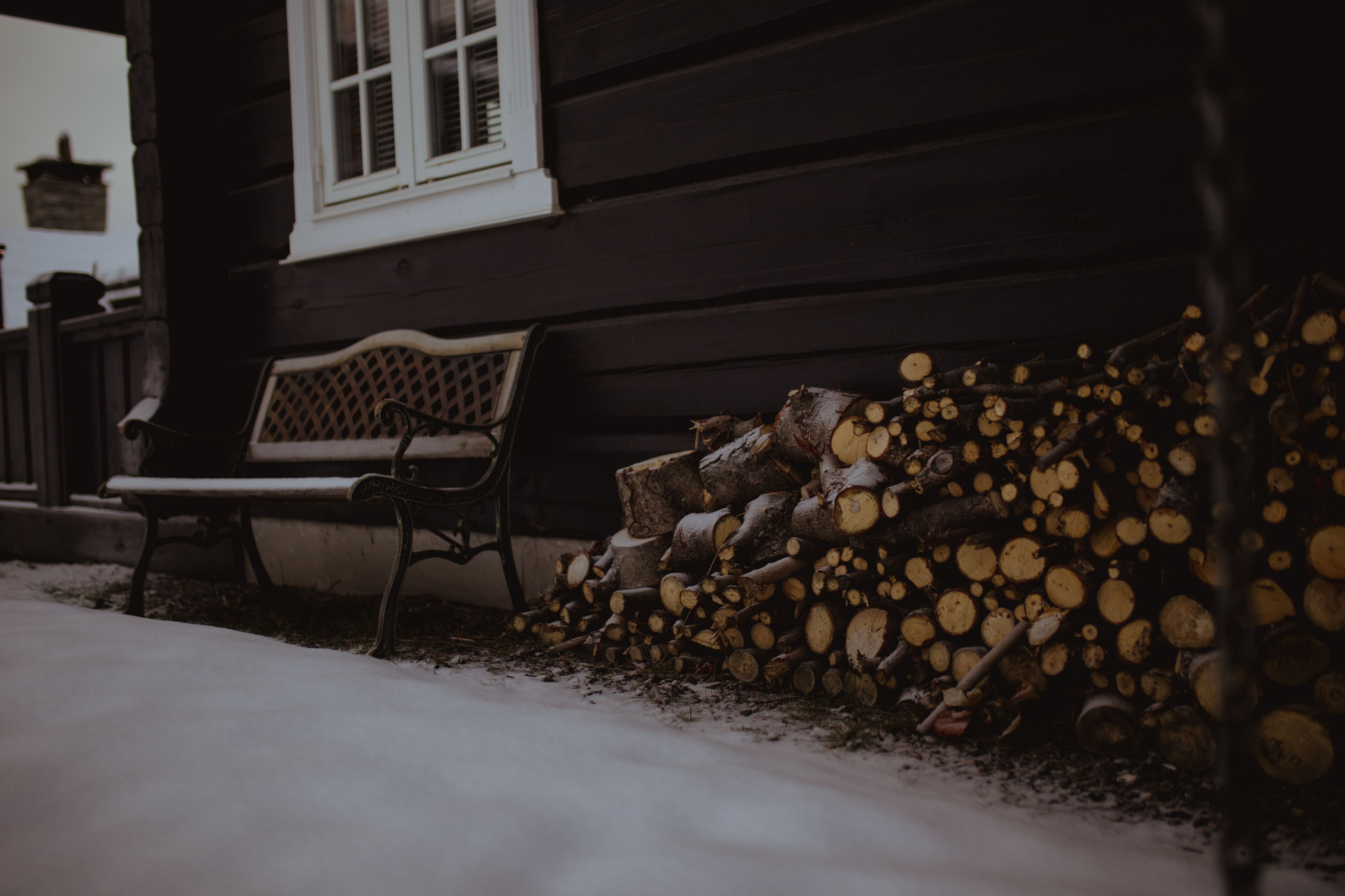 Snow-covered firewood stacked by a rustic house and empty bench, evoking winter coziness.