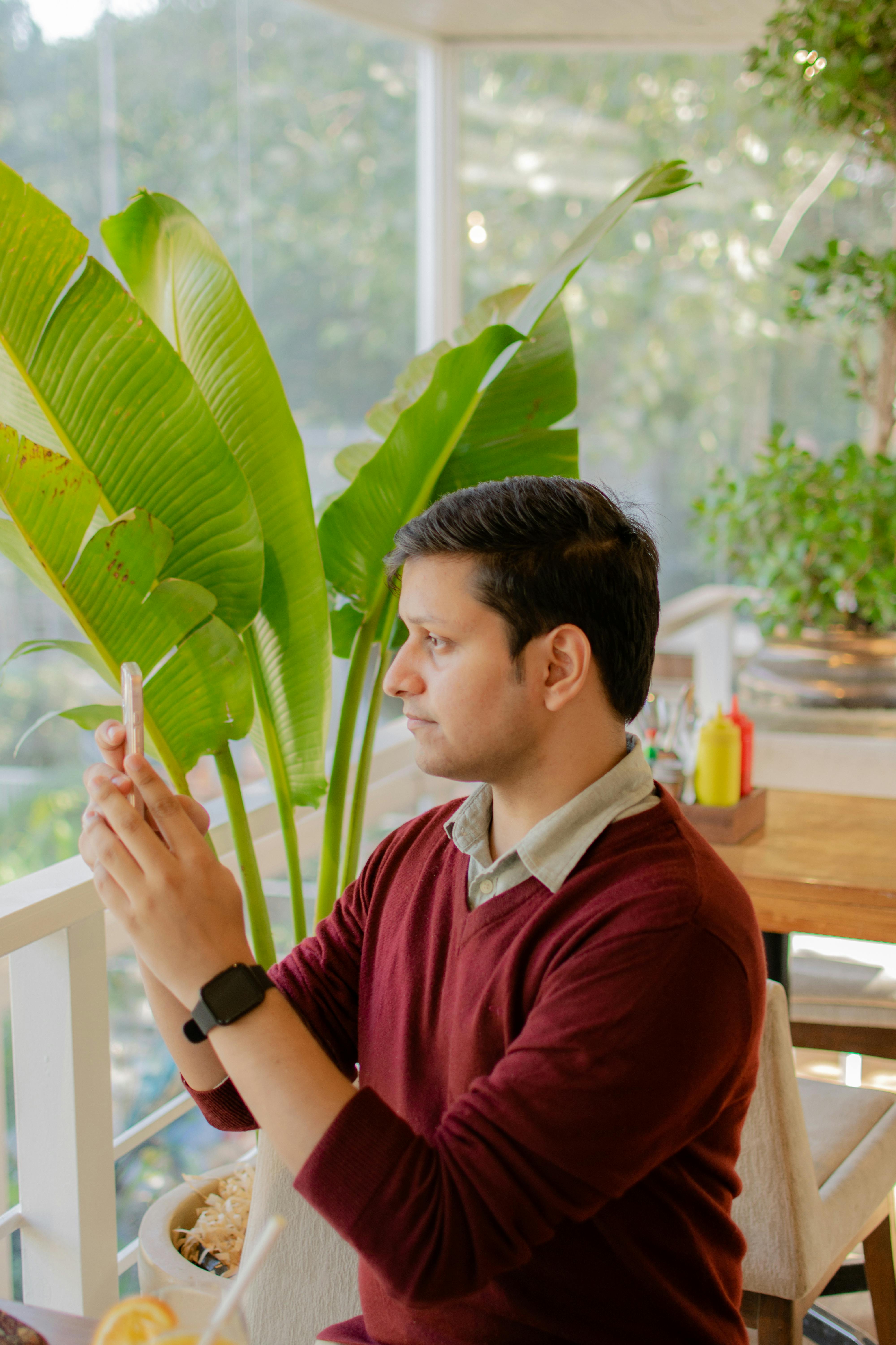 A man uses a smartphone to capture the scene in a modern restaurant with lush greenery and natural light.