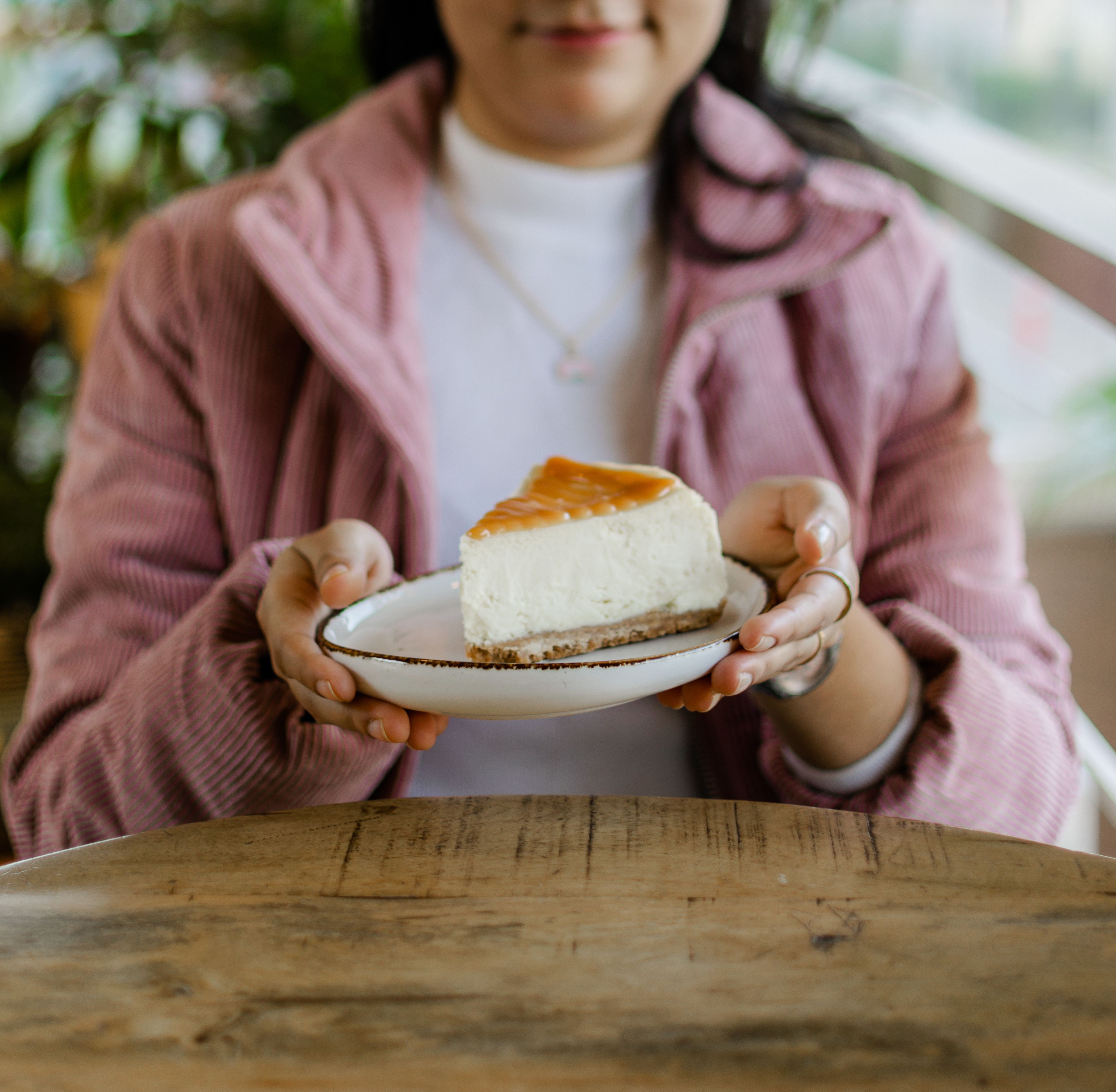 Portrait of a woman presenting a slice of cheesecake on a rustic ceramic plate.