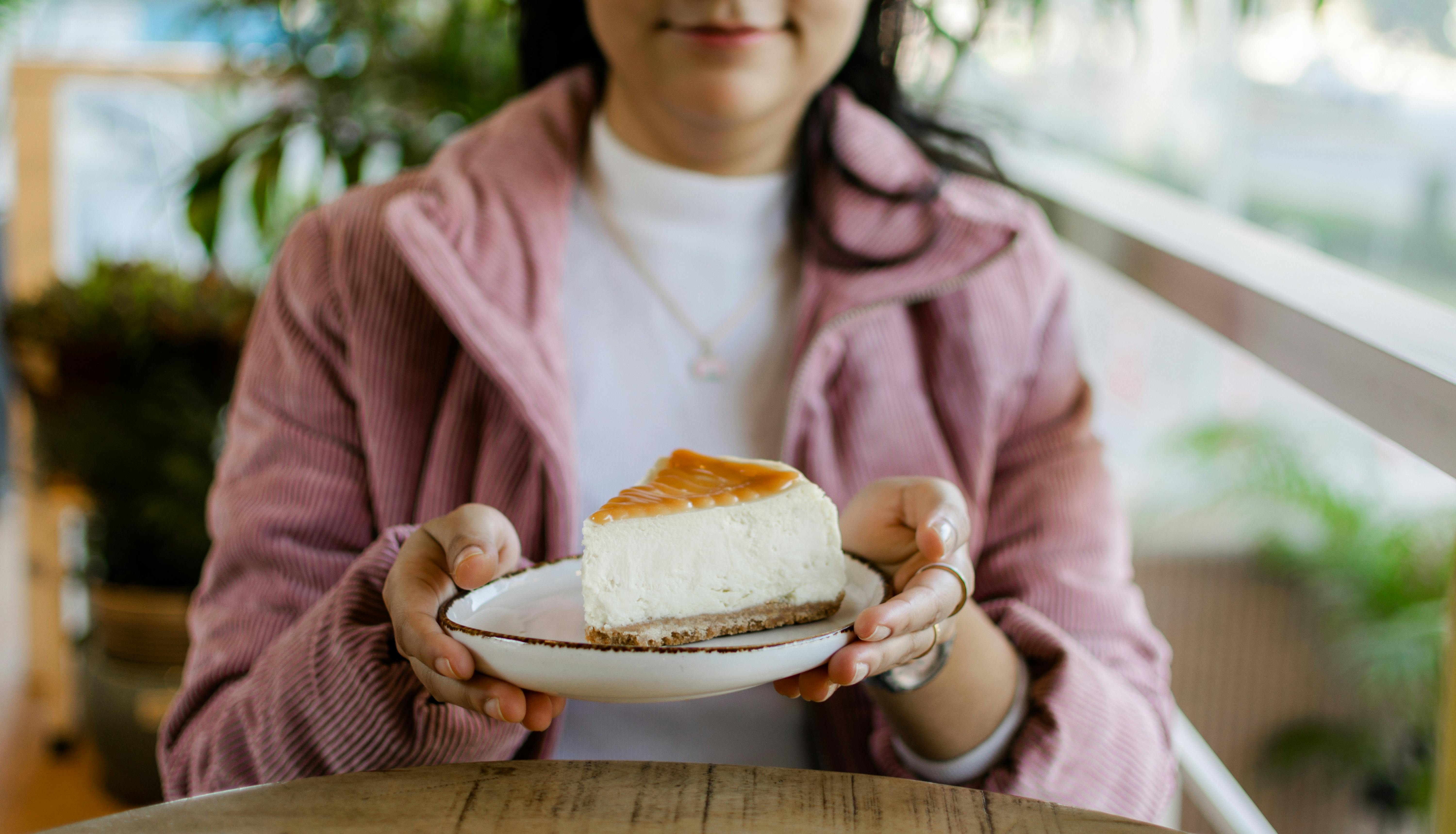 Woman holding a creamy cheesecake slice outdoors, perfect for dessert lovers.