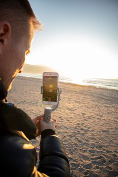 Man recording ocean sunset using a smartphone on the beach. Warm evening light enhances the scene.