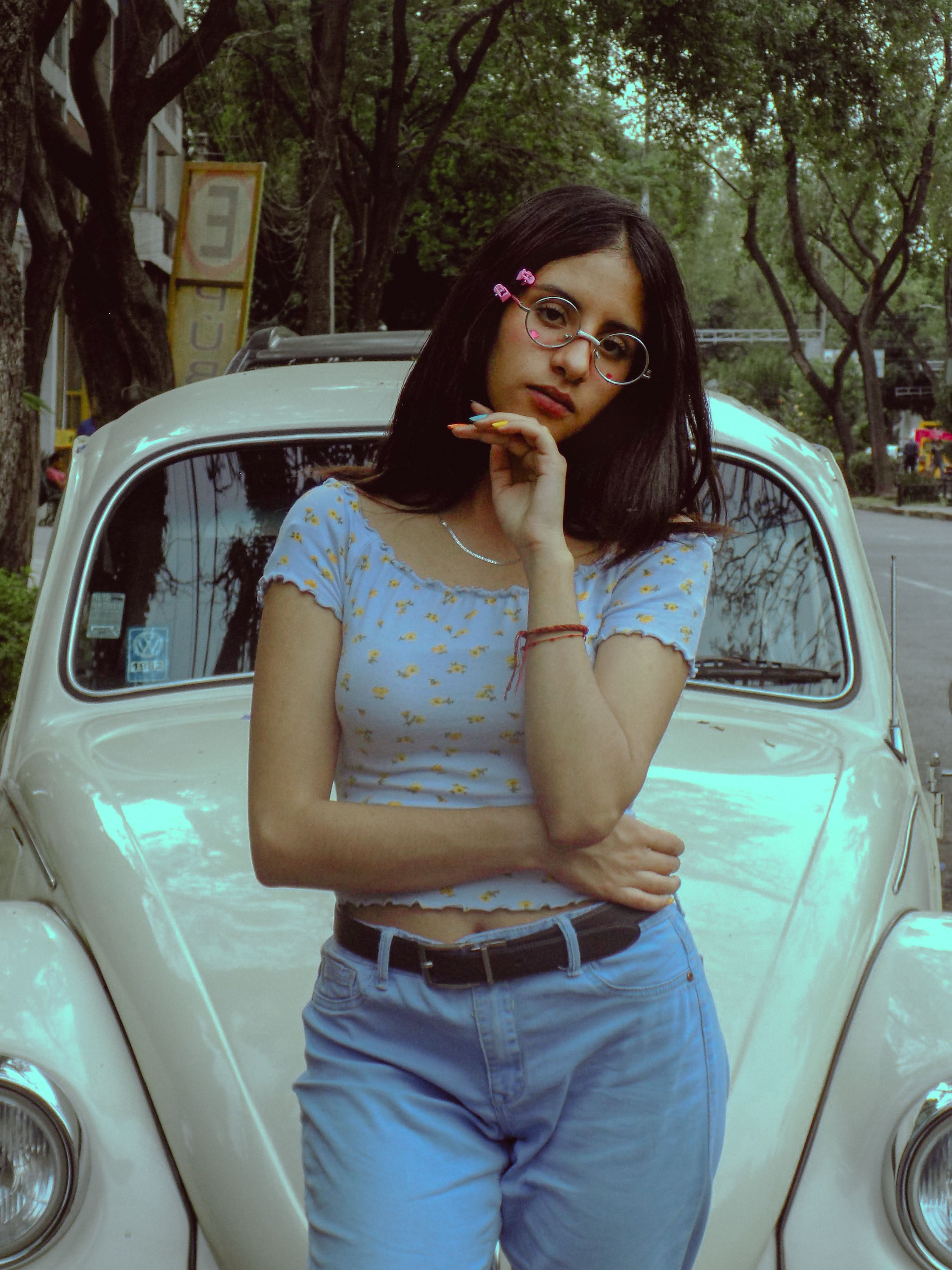 Young woman with glasses poses confidently by a vintage car, capturing classic outdoor style.