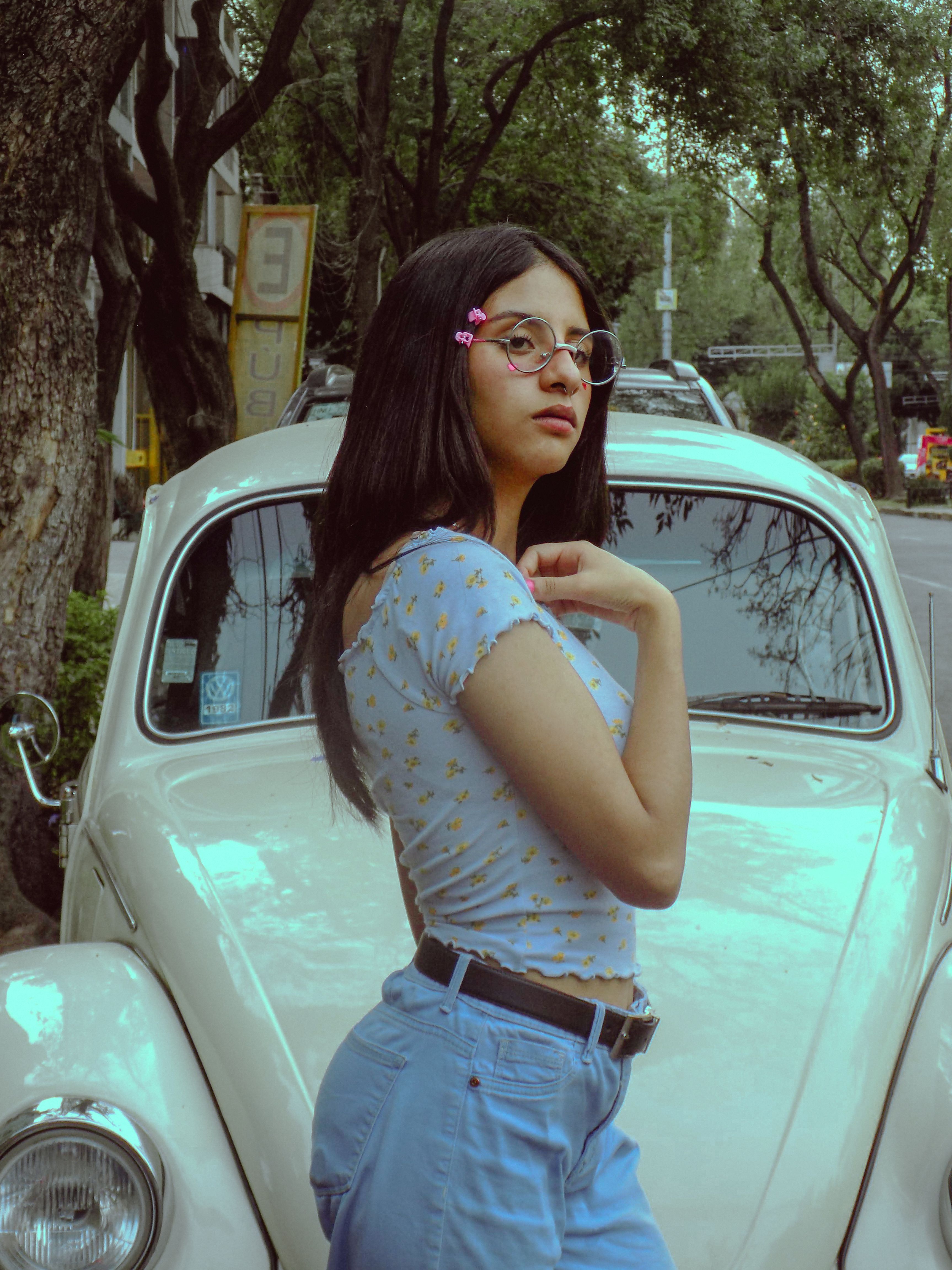 Stylish young woman in eyeglasses posing confidently beside a classic car on a city street.
