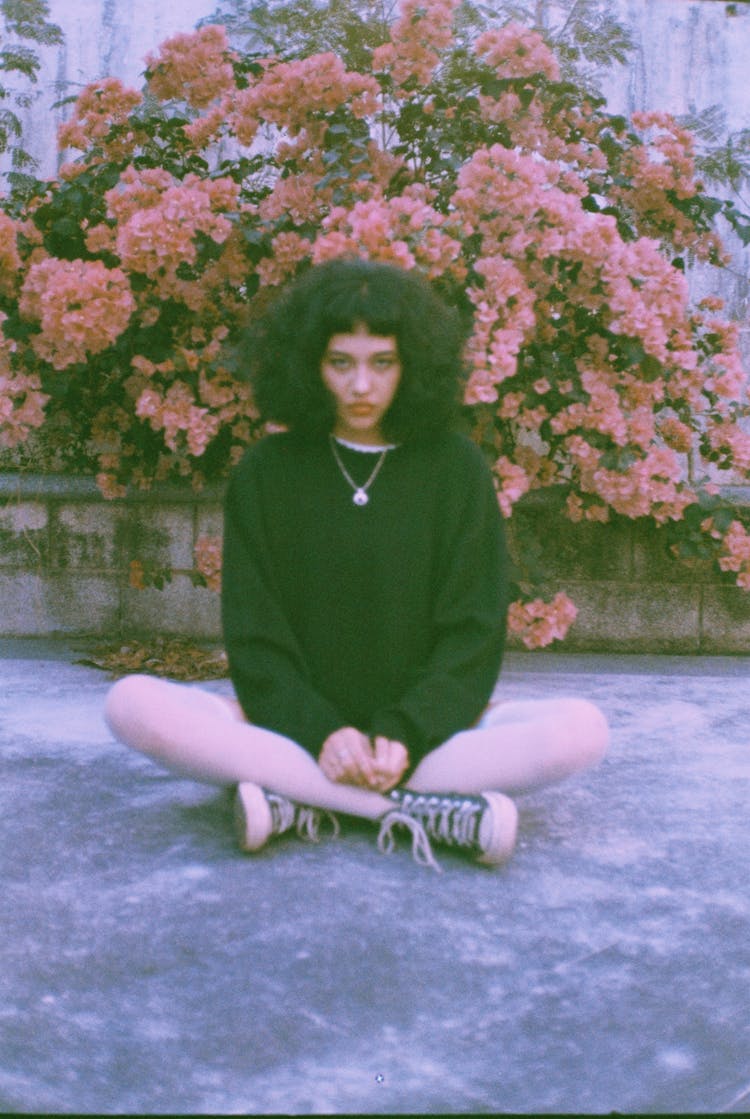 Photo Of A Young Woman Sitting With Crossing Legs In Front Of A Blossoming Bush