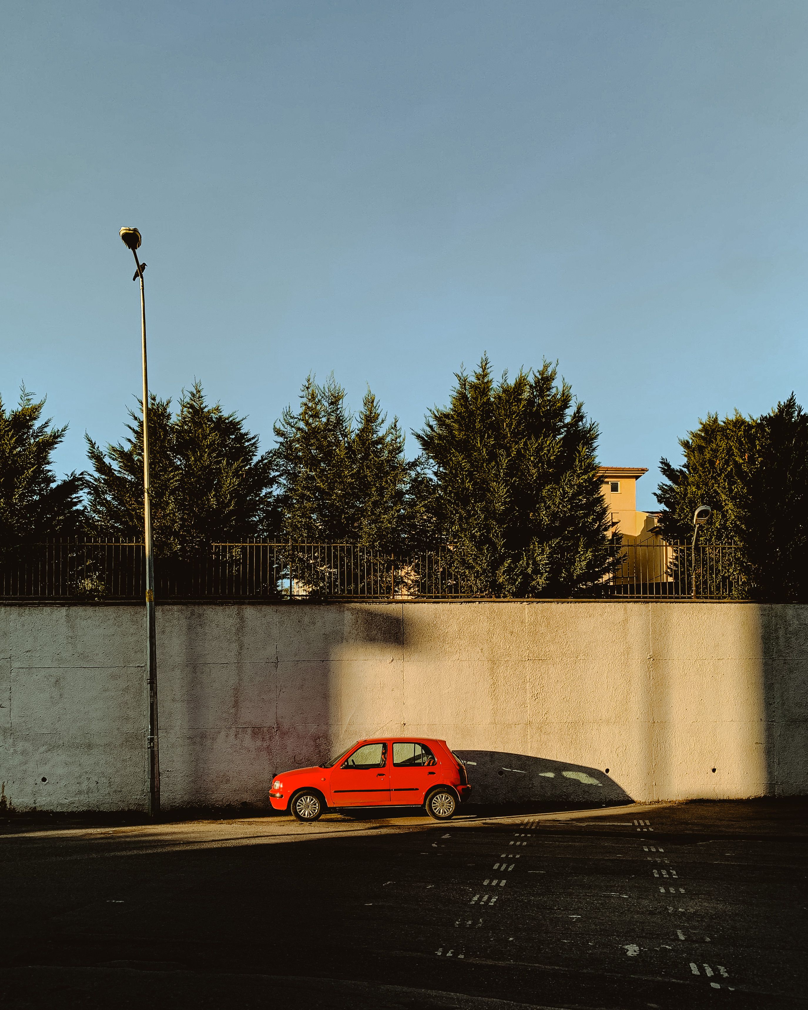 A red car parked by a concrete wall under bright sunlight on a summer day, casting a strong shadow.