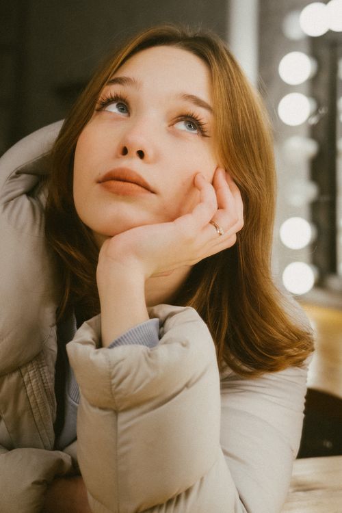 Free A young woman in a cozy puffer jacket looks up thoughtfully indoors, creating a warm and introspective mood. Stock Photo