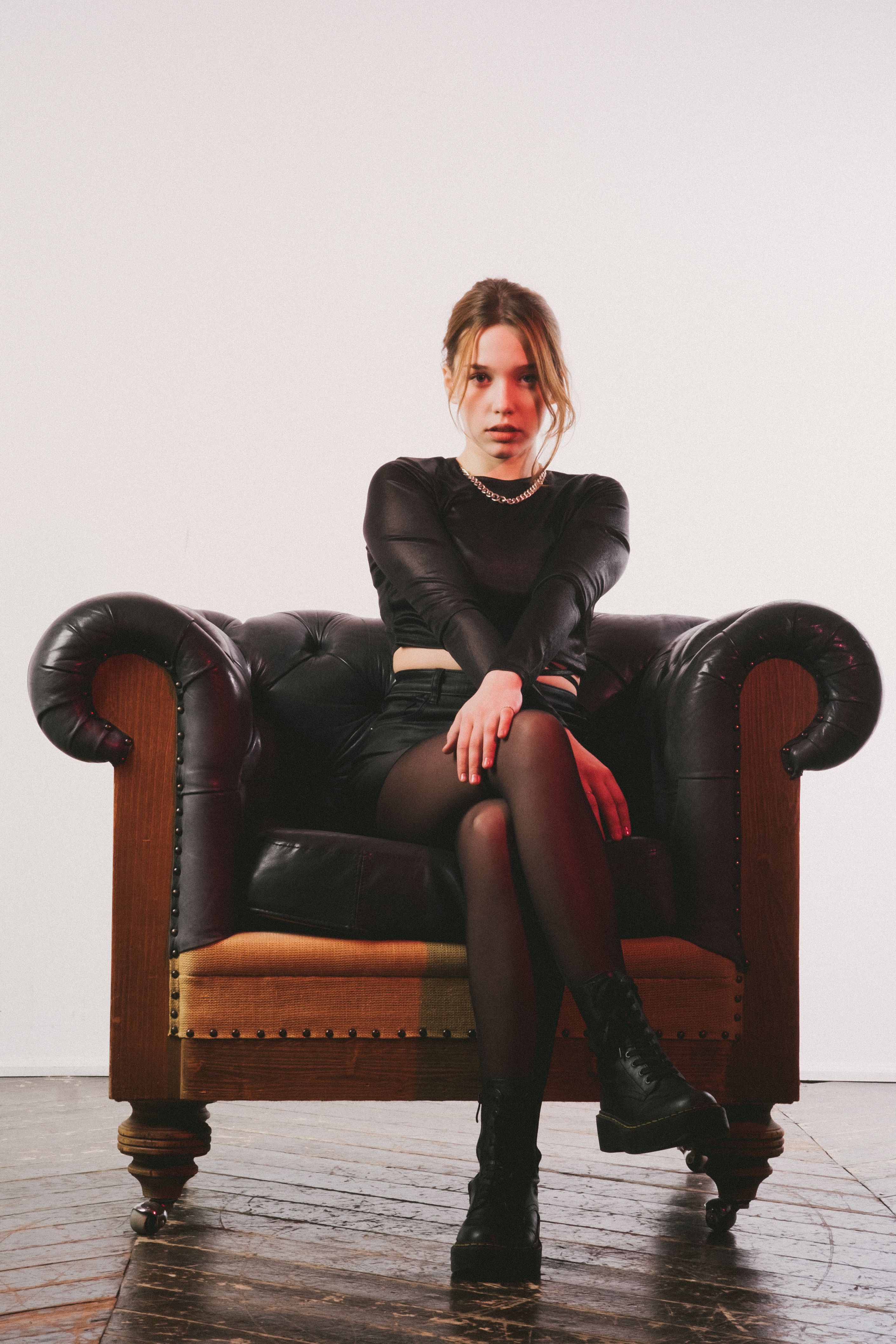 Fashionable woman in black attire sits confidently on a vintage leather armchair indoors.