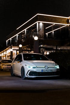 A Volkswagen Golf GTI parked on a city street at night, with illuminated lights and modern architecture.