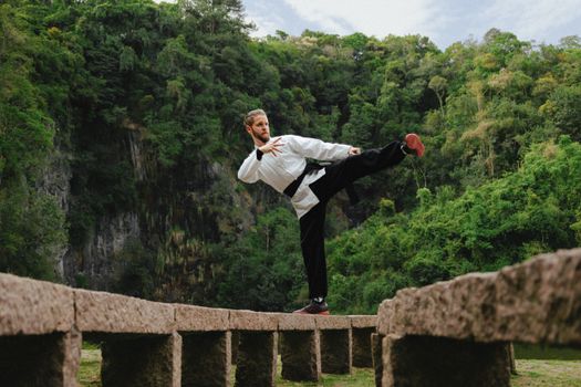 Martial artist in a white uniform balancing on a footbridge in a lush green valley, showcasing fitness and skill.