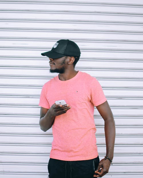 男人穿着粉红色的t恤，拿着智能手机站在灰色的卷起门旁边