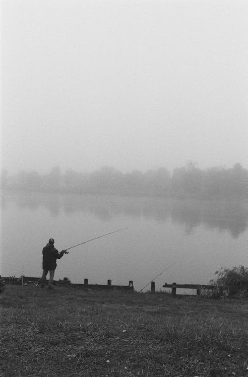 Grayscale Photo of a Person Standing on Grass Field while Fishing on the Lake