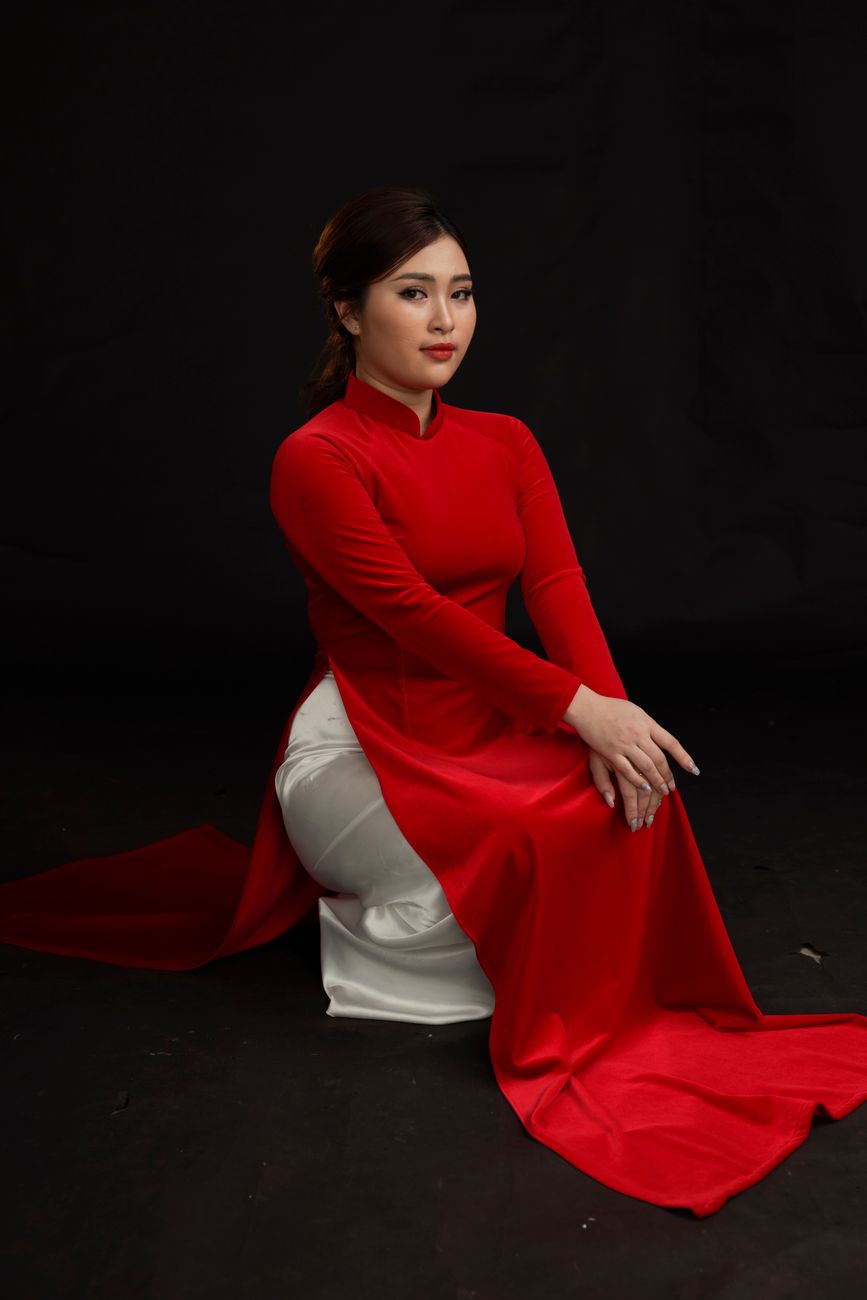 Portrait of an Asian woman in red ao dai dress, seated against a dark background, showcasing traditional elegance.