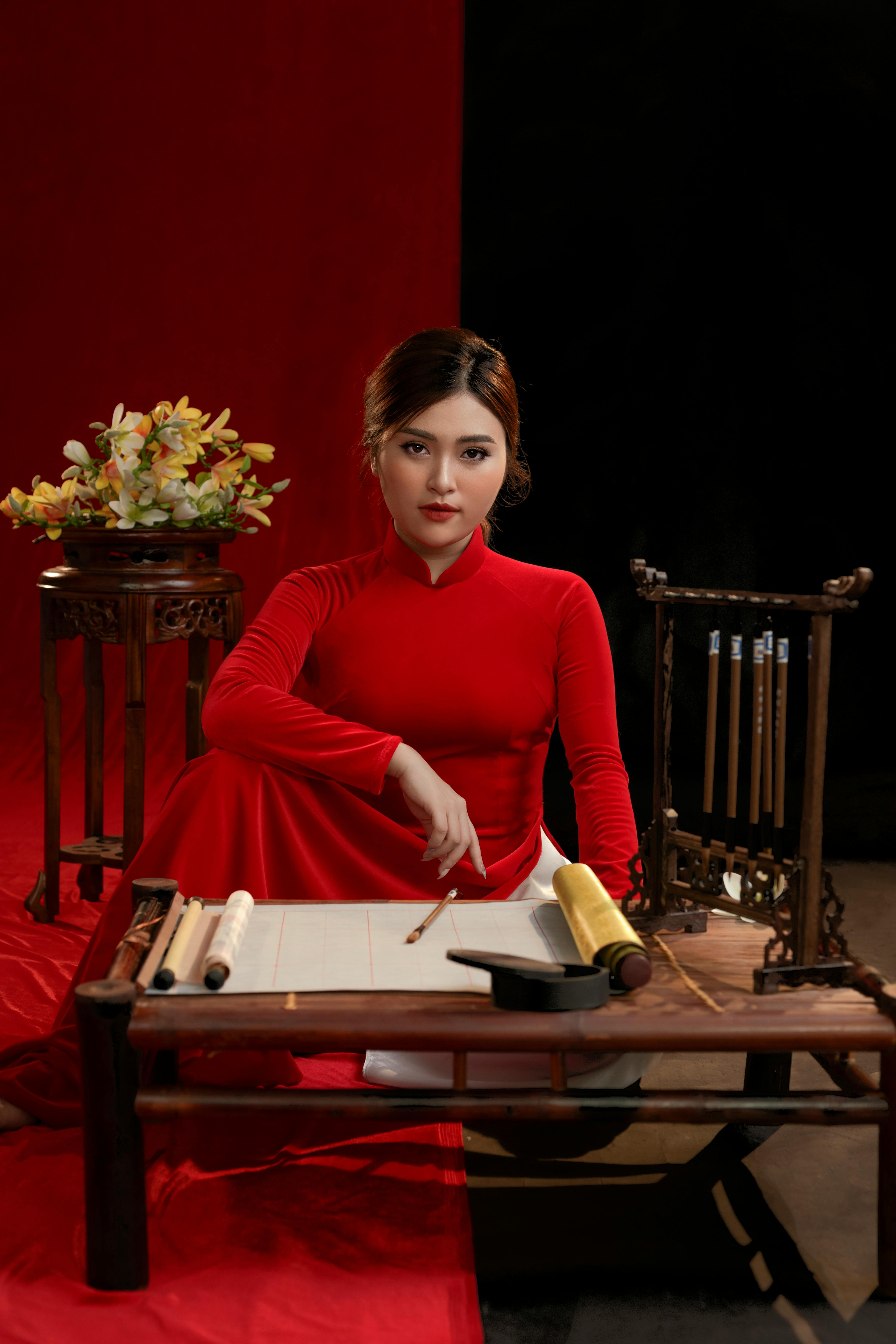 Asian woman in red ao dai poses in a studio with traditional decor.
