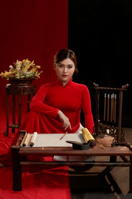 Asian woman in red ao dai poses in a studio with traditional decor.