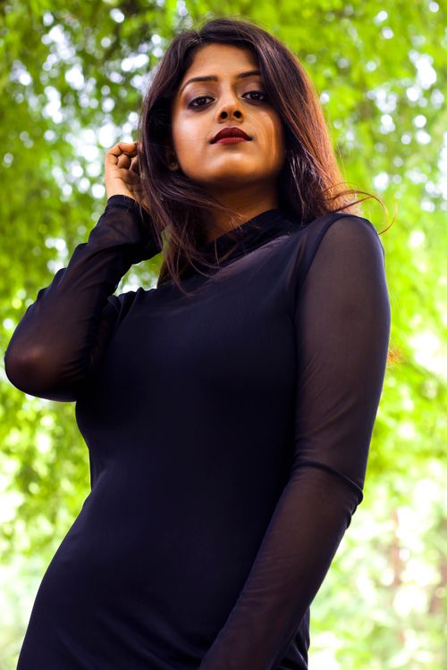 Free Elegant young woman in a black dress posing confidently outdoors with lush green foliage in the background. Stock Photo