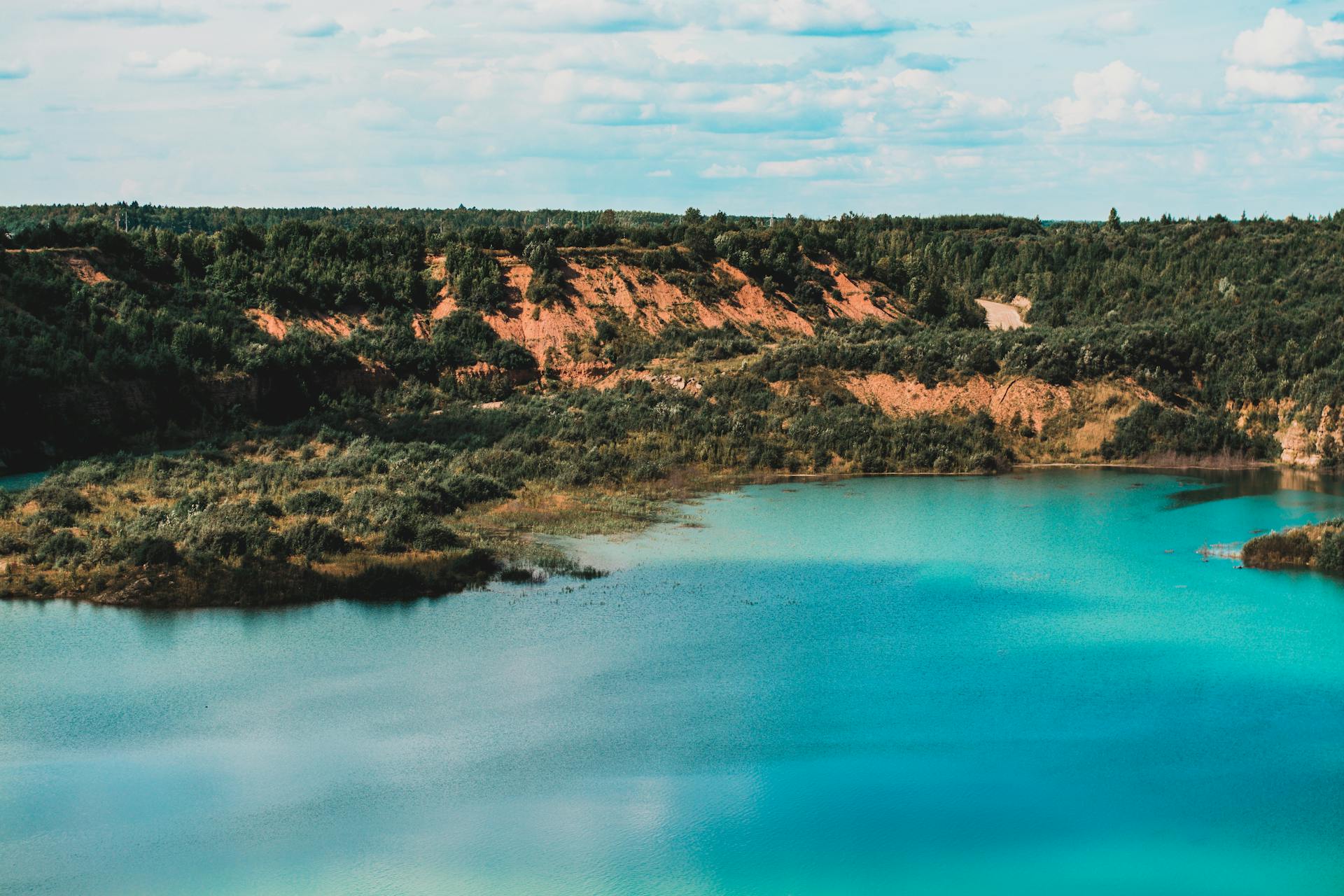 A scenic aerial view of an azure lake surrounded by lush green forest and hills under a bright blue sky.