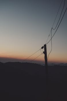 A tranquil scene of a silhouetted power line against a dusky sky over rolling hills at twilight.