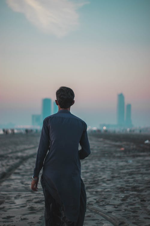 Free Adult male walking on a beach at dusk with a city skyline in the distance. Stock Photo
