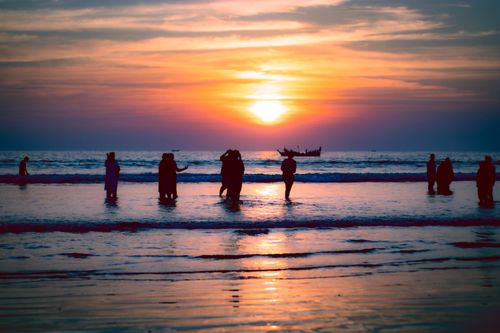Free Silhouetted people enjoy a serene sunset on the ocean shore, capturing vibrant hues and a calm sea. Stock Photo