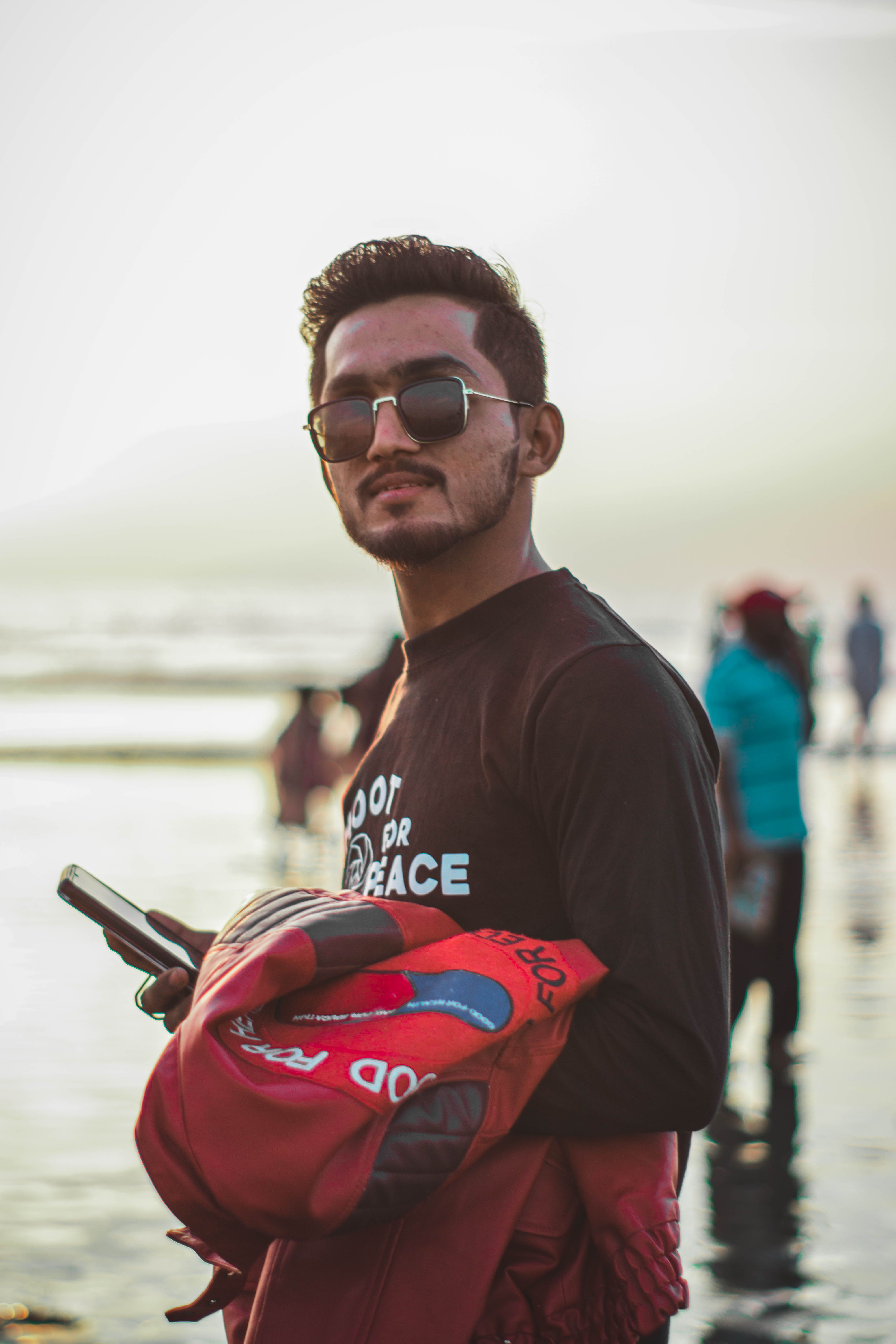 Young man wearing sunglasses and holding a jacket on a beach during sunset.