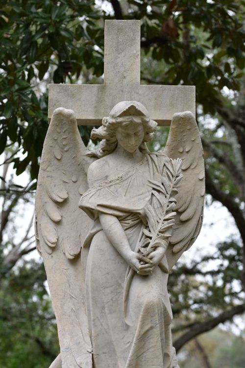 Free Graceful stone angel statue with a cross backdrop in a serene outdoor cemetery setting. Stock Photo
