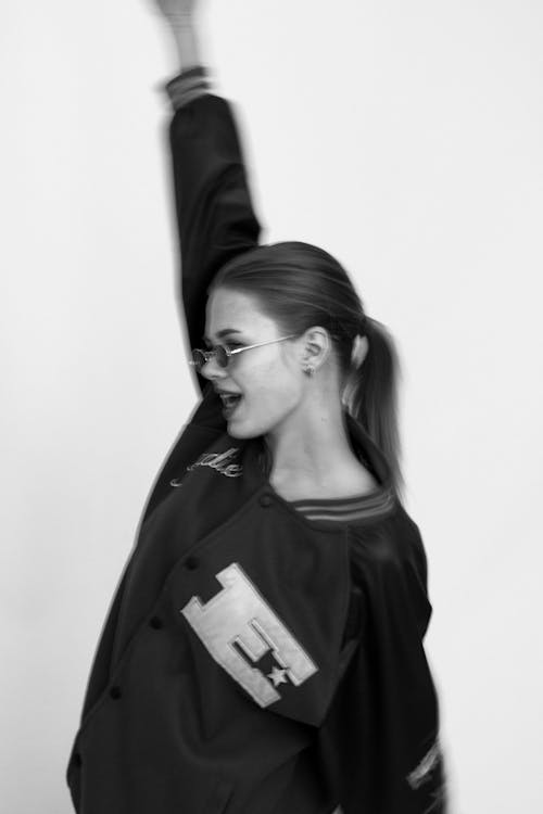 Grayscale Photo of Woman in Varsity Jacket Raising Her Hand 
