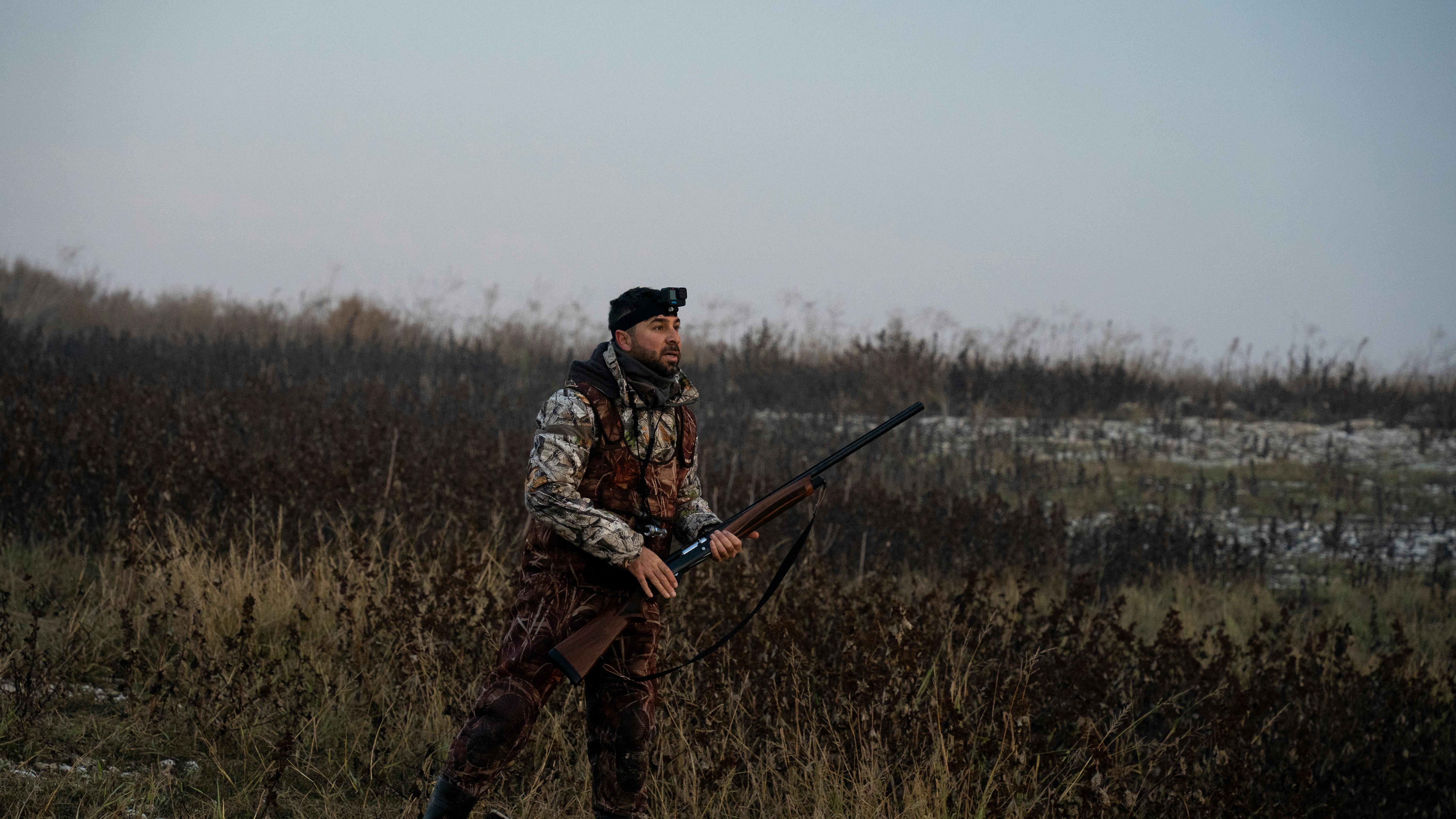 A male hunter in camouflage holds a rifle while traversing a misty field in early morning light.