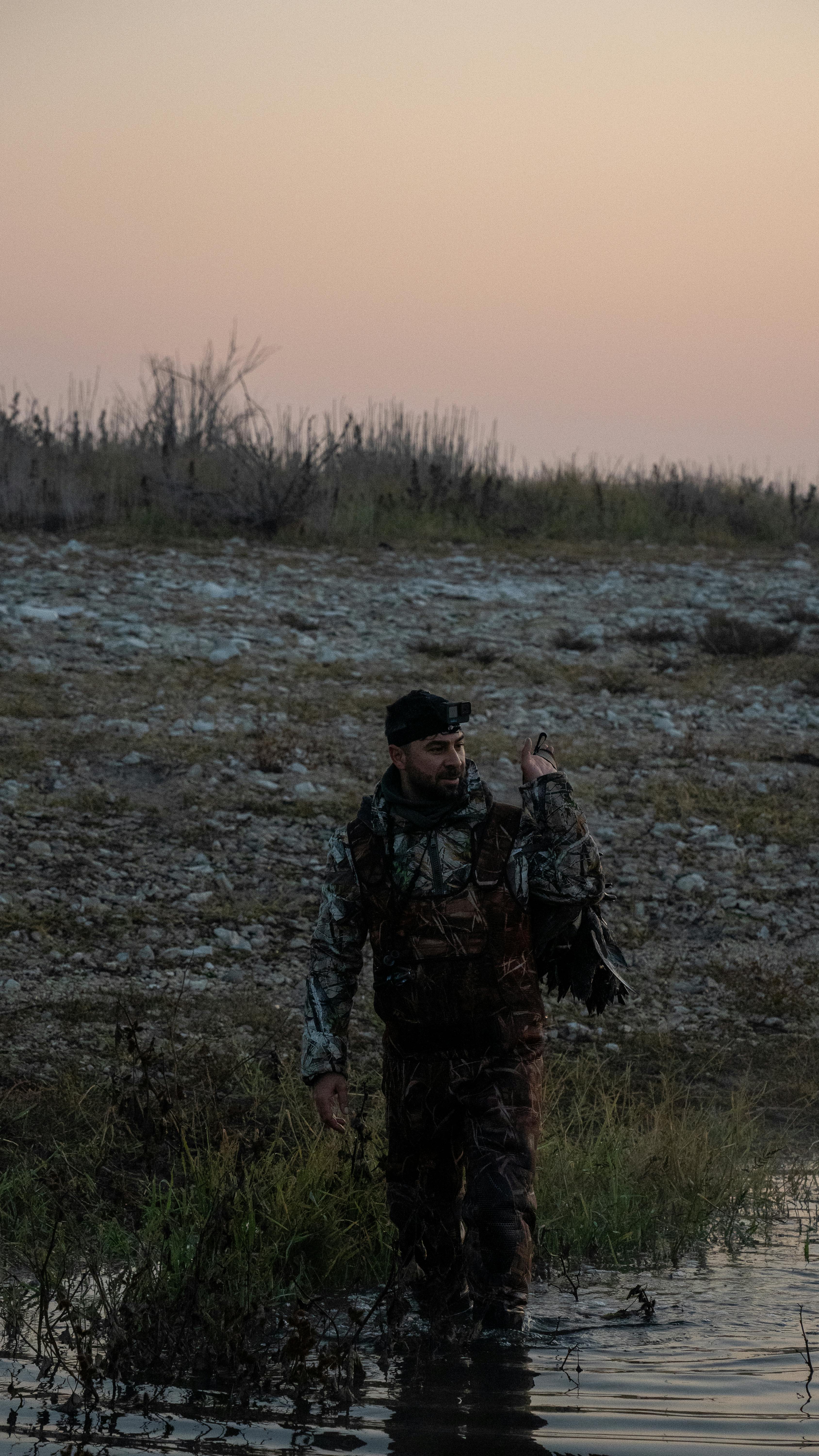 A man in camouflage stands by a lakeshore during dusk, carrying hunting gear.