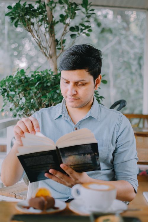 Free A young man enjoys reading a novel in a cozy indoor cafe setting next to greenery. Stock Photo