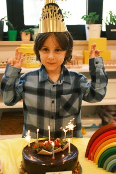 A young boy celebrates his birthday with a cake, candles, and party hat indoors.