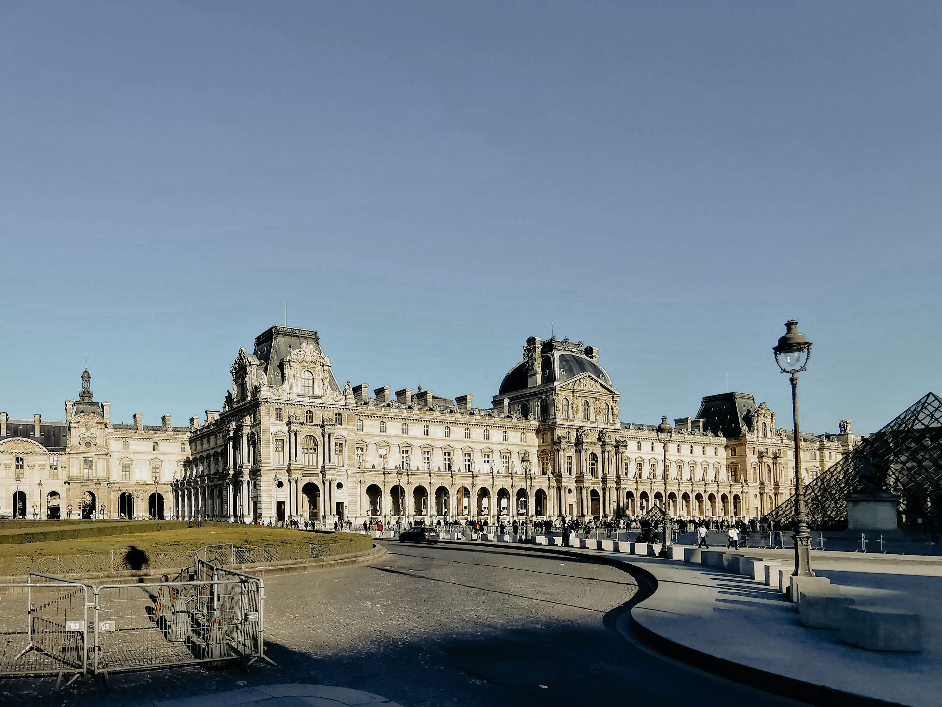 Sunlit Louvre Museum in Paris, France, with ornate palace façades, glass pyramid, courtyard gardens, and visitors under a blue sky