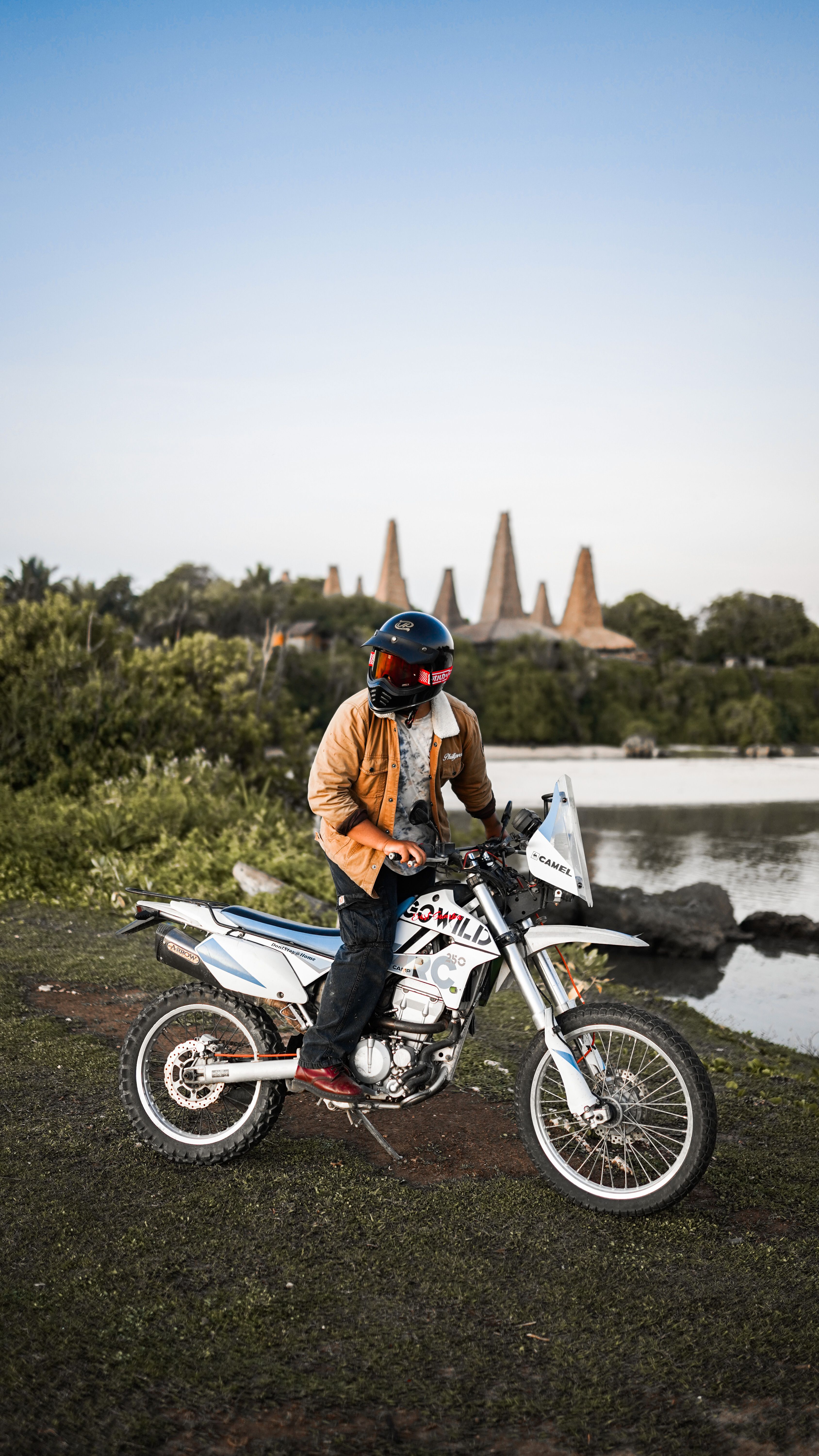 Man on a motorcycle by a serene lakeside with unique architecture in the background. Adventure and exploration.