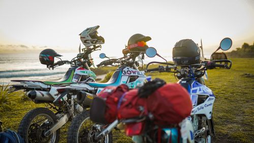 Free Motorbikes lined up on a grassy shore as the sun sets by the ocean, ready for adventure. Stock Photo