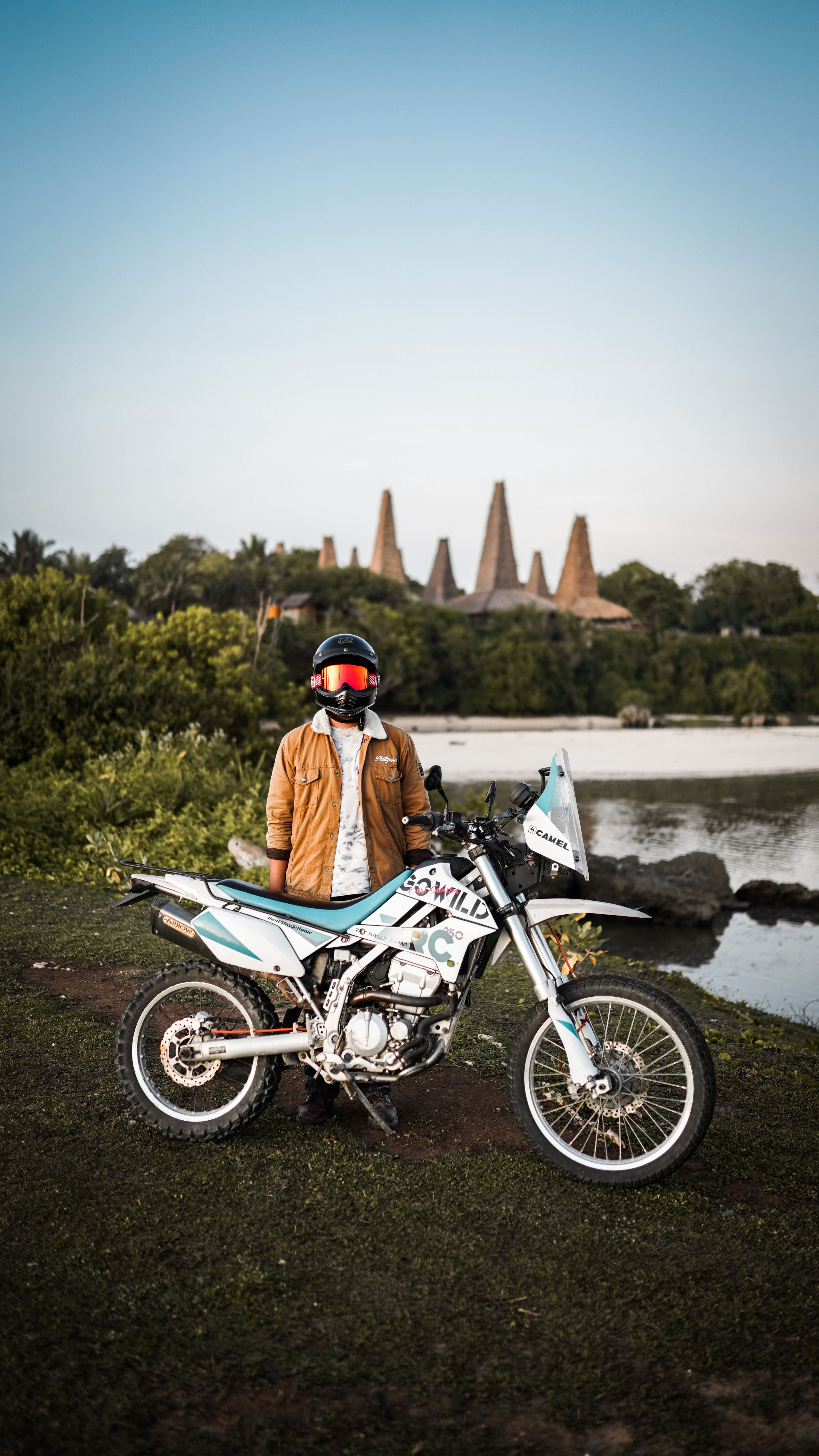 A person in a brown jacket and helmet standing beside a dirt bike near a scenic water body.