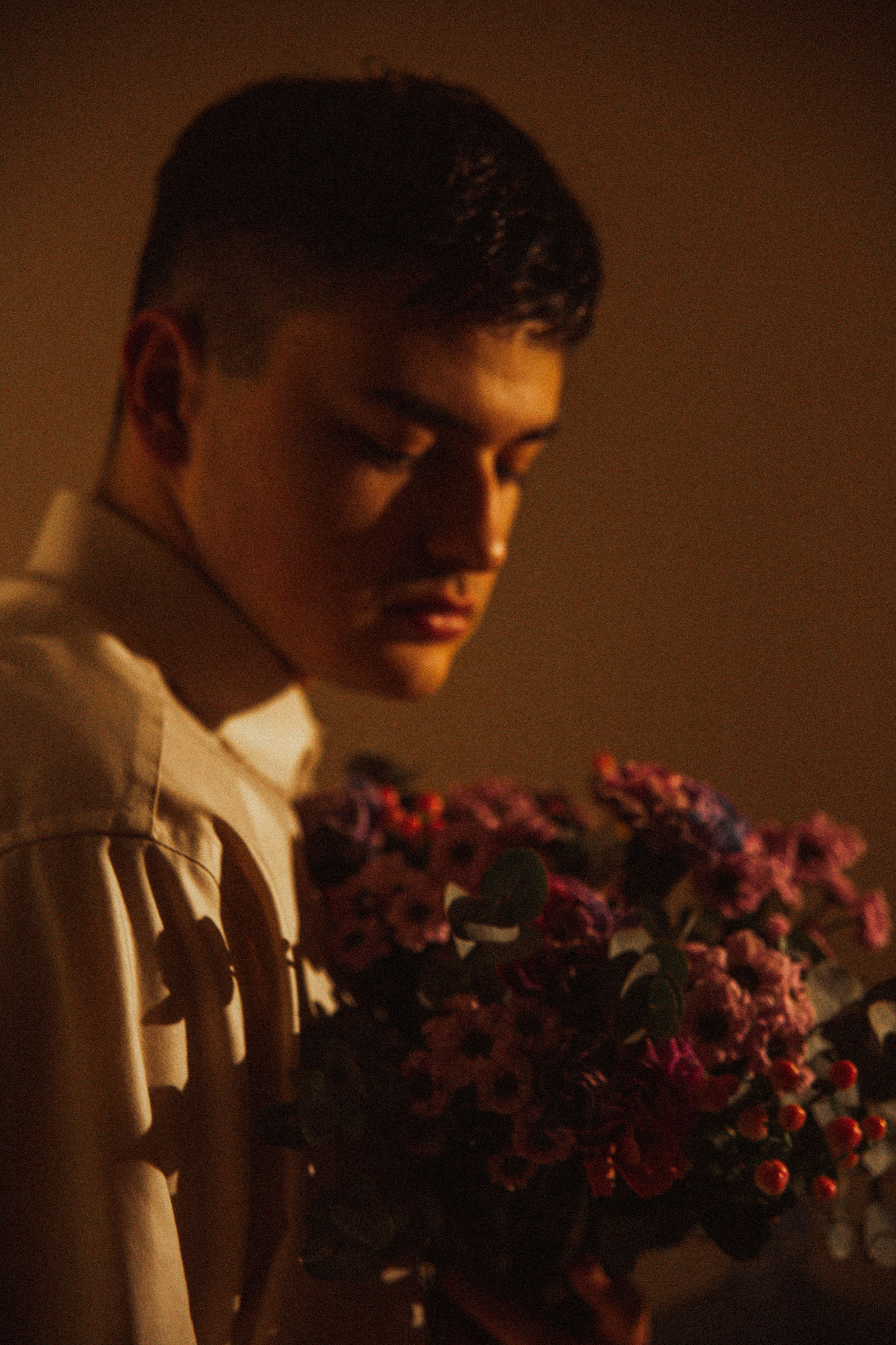 A young man gazes thoughtfully at a bouquet of flowers in soft, warm lighting.