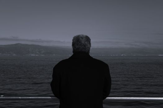 A solitary man gazes at the vast ocean from a pier, captured in dramatic grayscale.